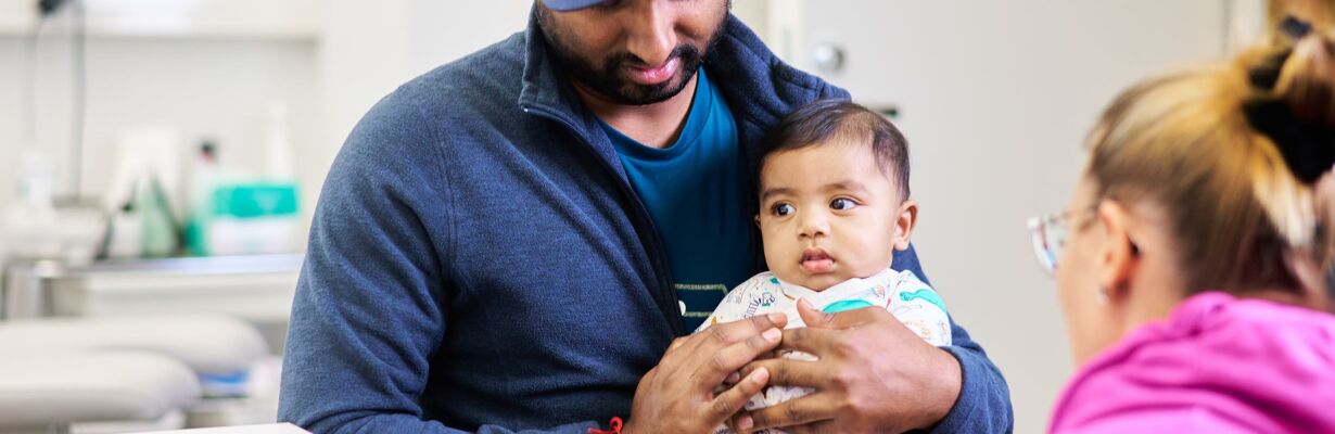 Kuvassa maahanmuuttajataustainen isä vauvansa kanssa neuvolassa, hoitaja antaa vauvalle rokotteen