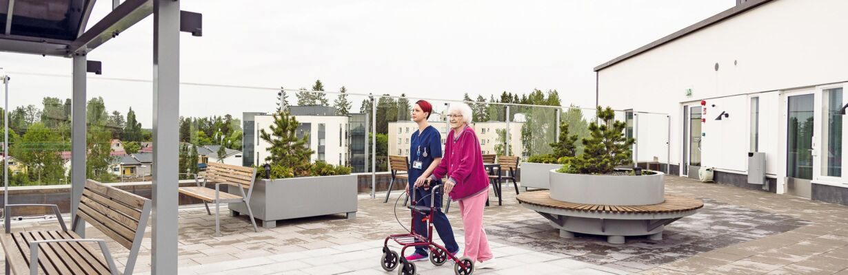 Hoitaja ulkoiluttaa rollaattorin kanssa kulkevaa ikäihmistä.
