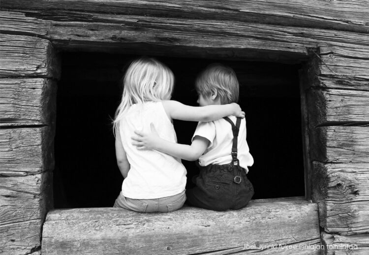 Kaksi lasta istuvat selin ikkunalaudalla, halaten toisiaan.