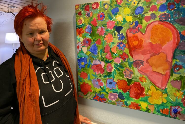 Taiteilija Eveliina Narkilahti maalaamansa taideteoksen vieressä. Taideteos on värikäs taulu, jossa on iso punainen sydän ja paljon erivärisiä kukkia.