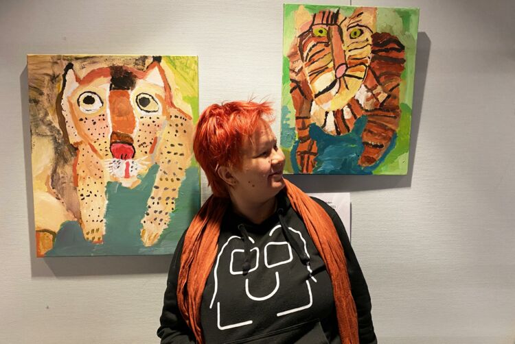 Taiteilija Eveliina Narkilahti maalaamiensa taideteosten vieressä. Taideteokset ovat tauluja, jotka esittävät kissaeläimiä.