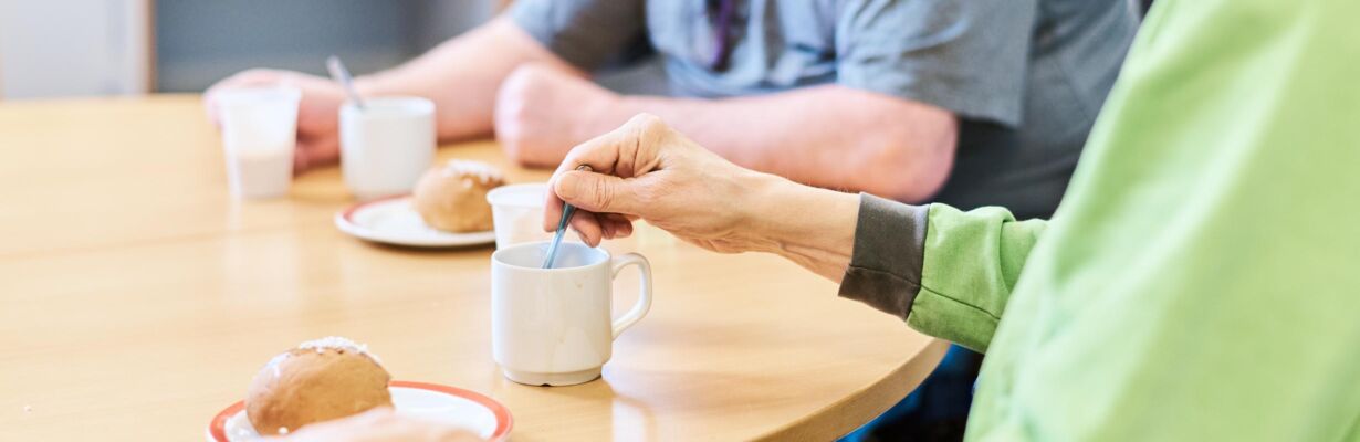 Kuvassa kaksi miespuolista henkilöä, jotka käyttävät pyörätuolia. Henkilöt istuvat kahvipöydän ääressä, juovat kahvia ja syövät pullaa.