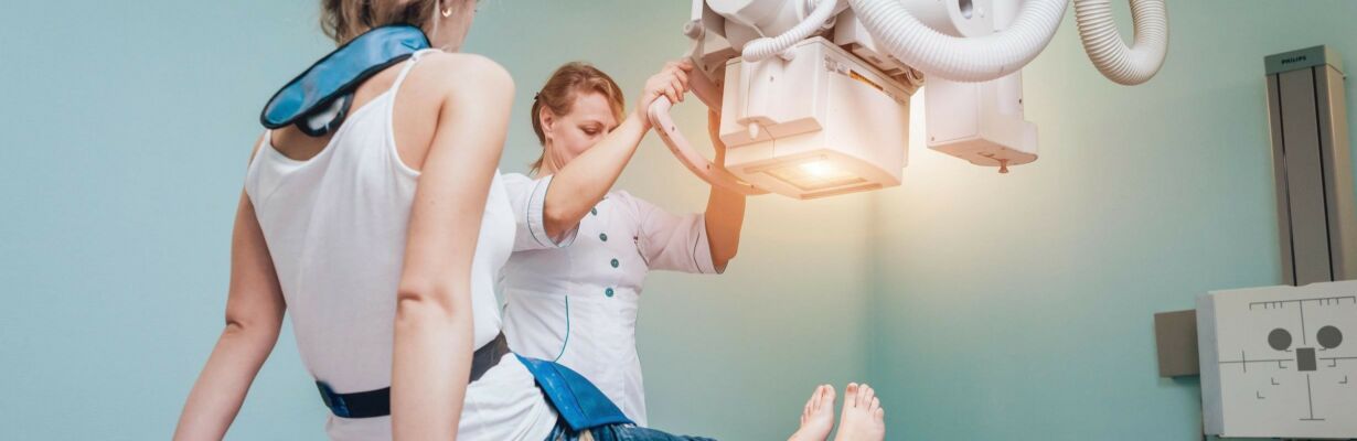 Kuvassa otetaan röntgenkuvat nuoren naisen jaloista. Työntekijä säätää laitetta.