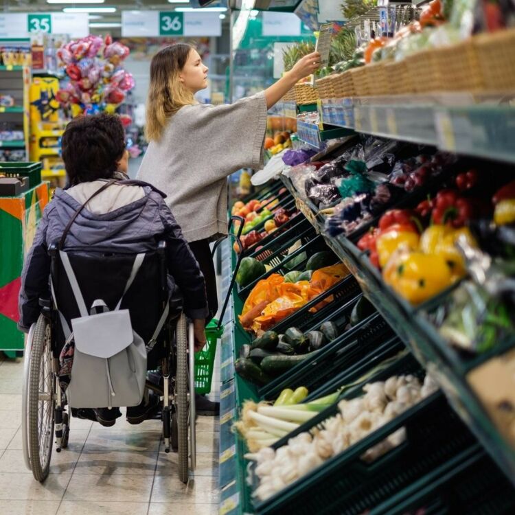 Kuvassa nuori nainen avustaa pyörätuolilla liikkuvaa henkilöä ruokakaupassa ostosten tekemisessä.