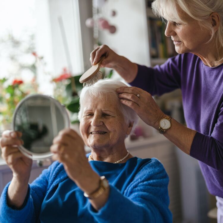 Kuvan naishenkilö kampaa ikääntyneen naisen hiuksia. Ikäihminen katsoo itseään peilistä.