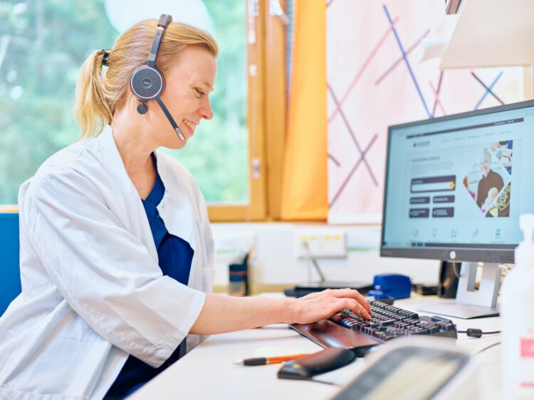 Keusoten lääkäri istuu toimistopöydän äärellä tietokoneella, hänellä on korvillaan headset-kuulokkeet.