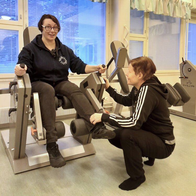 Kuvassa toimintaterapeutti Soile Sillman ja fysioterapeutti Satu Alvela näyttävät, miten jalkojen lihaksia voimistetaan kuntolaitteen avulla.