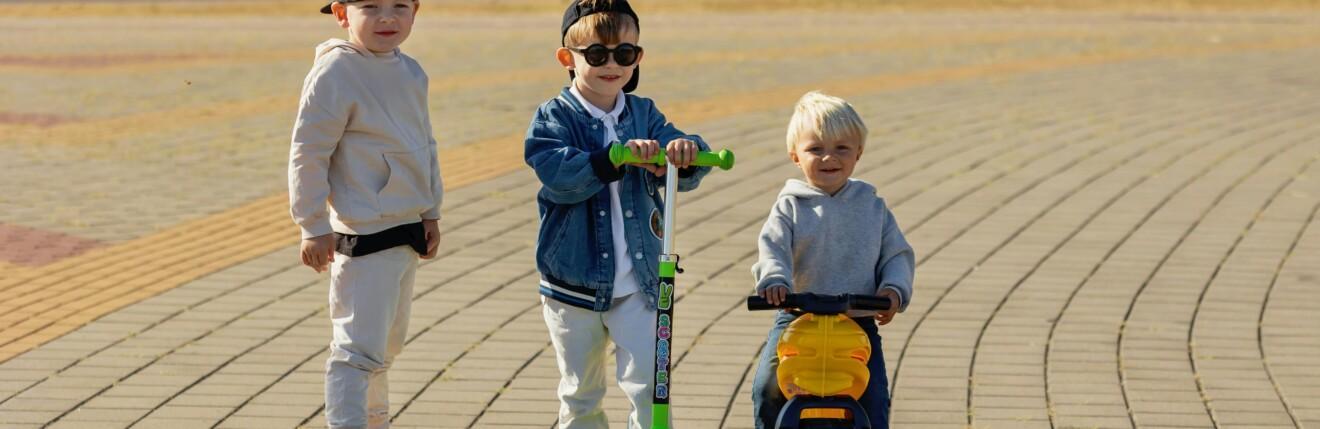 Kolme pikkulasta skeittilaudoilla ja leikkimopolla ulkona.
